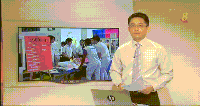Male news anchor in shirt and tie stands beside a TV screen showing people in a medical or classroom setting.